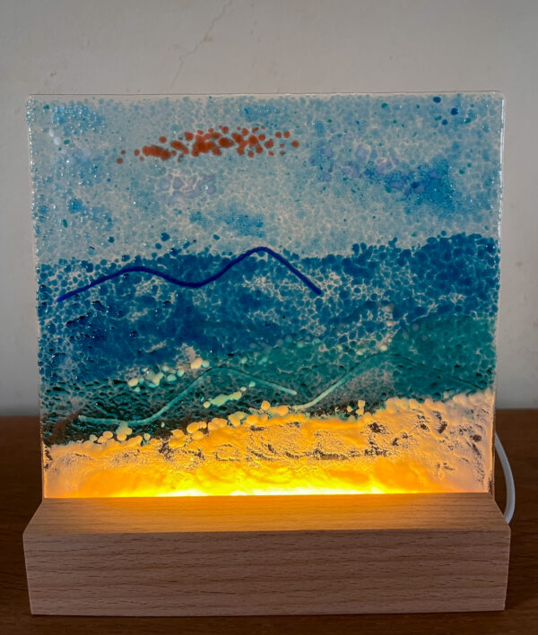 Een handgemaakt glazen creatie op een verlichte sokkel met als thema strand en zee.